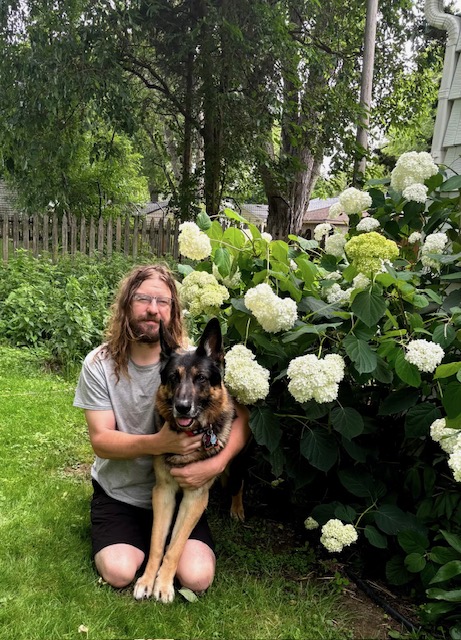 A portrait of Peter Schneider and his dog, next to a bush of white hydrangeas.